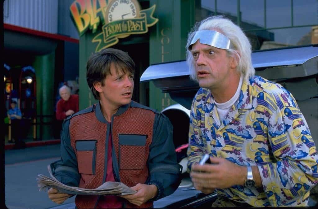 Marty McFly and Doc Brown together in the futuristic 2015 Hill Valley setting from Back to the Future Part II, both in their iconic outfits — Marty in the orange jacket and flat cap, Doc in his futuristic coat and sunglasses