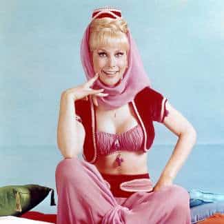 Jeannie I Dream of Jeannie cosplay reference showing the full character look, pink harem costume, short blonde wig styled into a high ponytail, mini fez hat, chiffon scarf, and genie bottle prop
