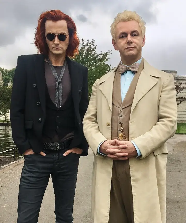 Crowley and Aziraphale from Good Omens shown together as a Halloween costume duo, with Crowley in his all-black outfit, long wool coat, and steampunk sunglasses alongside Aziraphale in his cream and fawn angel aesthetic, the most beloved group costume pairing from the Good Omens TV series