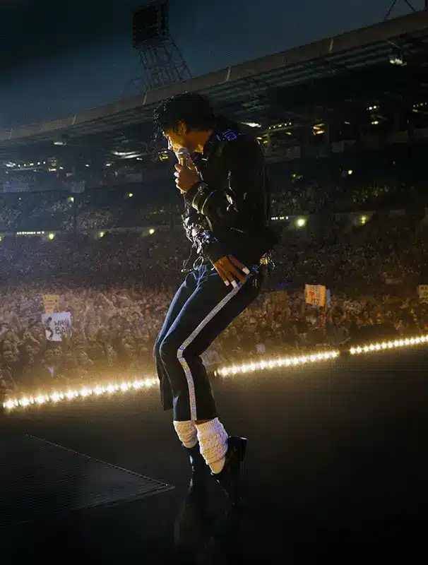 Jaafar Jackson as Michael Jackson performing on stage in the Michael 2026 biopic, wearing the striped trousers, belted leather jacket, curly wig, and holding a white microphone