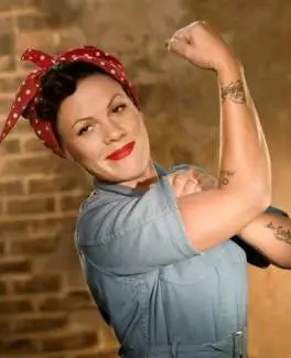 P!nk poses as Rosie the Riveter in a light blue work shirt and red polka dot bandana, rolling up her sleeve and flexing against a brick wall