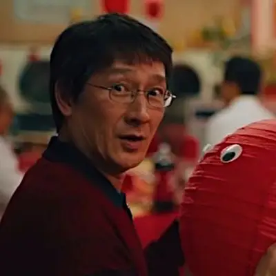 Waymond Wang wearing wire-rimmed glasses and a dark top, looking over his shoulder with a surprised expression beside a large red paper lantern with googly eyes in a laundromat setting