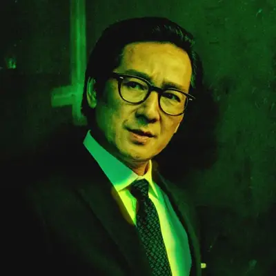 Waymond Wang in a black suit jacket, white collared shirt, and dark patterned tie with thick black-rimmed glasses, lit in green light against a dark background