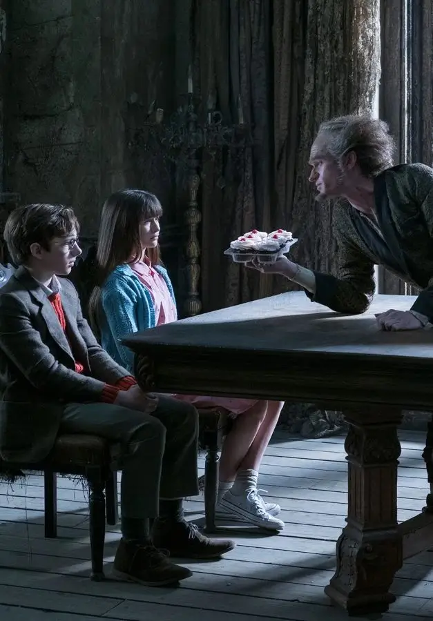 Count Olaf leans across a table offering cupcakes to Klaus and Violet Baudelaire in A Series of Unfortunate Events, used as a group Halloween costume reference