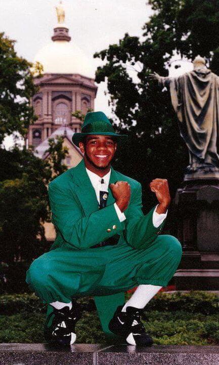 Student performer in the Notre Dame Fighting Irish Leprechaun mascot costume at a sporting event, showing the full green tailcoat, hat, and gold shoes
