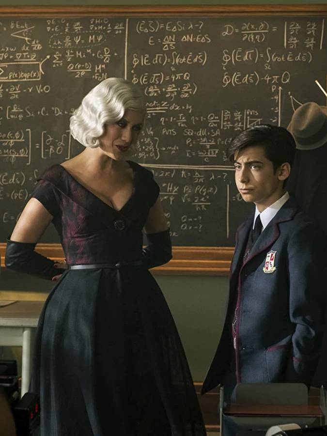 The Handler and Number Five from The Umbrella Academy facing each other, showing the visual contrast between her vintage leather trench coat and structured accessories and his school uniform, useful as a Halloween couples costume reference