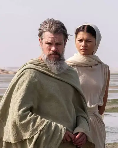 An older, weathered Odysseus wrapped in an olive-green cloak standing near the water with a woman in draped sand-colored clothing beside him