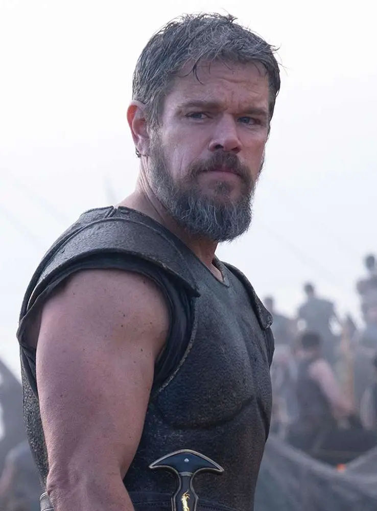 Matt Damon as Odysseus in The Odyssey wearing dark chest armor with a dagger at his waist, looking back over his shoulder with a weathered, dirt-streaked expression