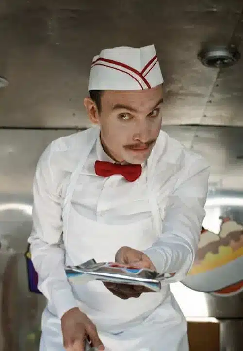 Jonathan Smiley the Ice Cream Man standing inside his ice cream truck in white vendor uniform with red bow tie