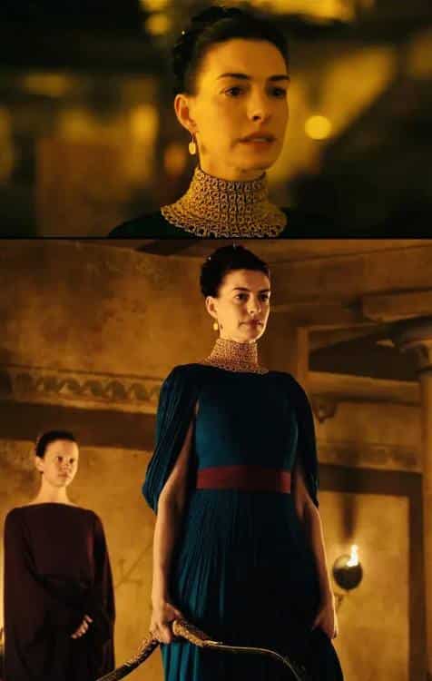 Anne Hathaway as Penelope in The Odyssey 2026 wearing a teal draped gown with gold mesh choker and burgundy sash, standing in a torch-lit palace hall