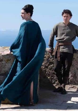 Penelope in a flowing teal draped gown seen from the side, standing near another character on a coastal stone rampart in The Odyssey 2026