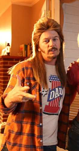 Joe Dirt wearing a red plaid sleeveless flannel shirt over a Pantera tank top with a mullet wig