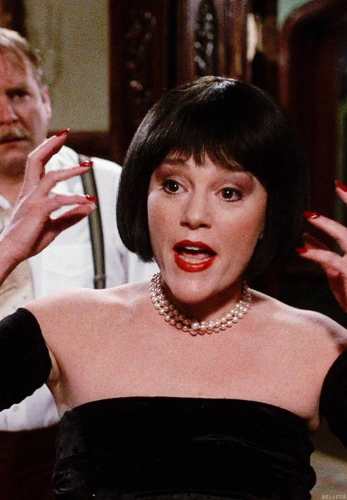 Mrs. White from Clue 1985 wearing a black dress with a veiled pillbox hat and bob wig