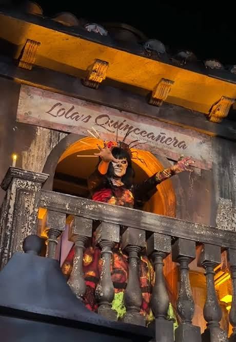 Lola from Killceañera performing on the balcony of her burned home at Halloween Horror Nights 2026, wearing the burgundy lace gown and gold sunburst crown