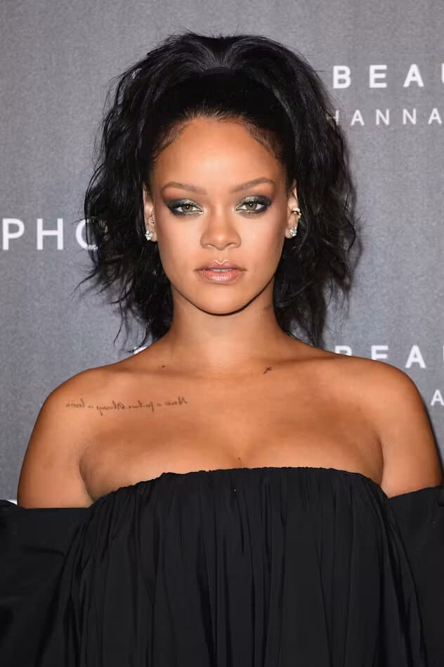 Rihanna in a black off-the-shoulder top at a beauty event, with the small script tattoo under her right collarbone visible